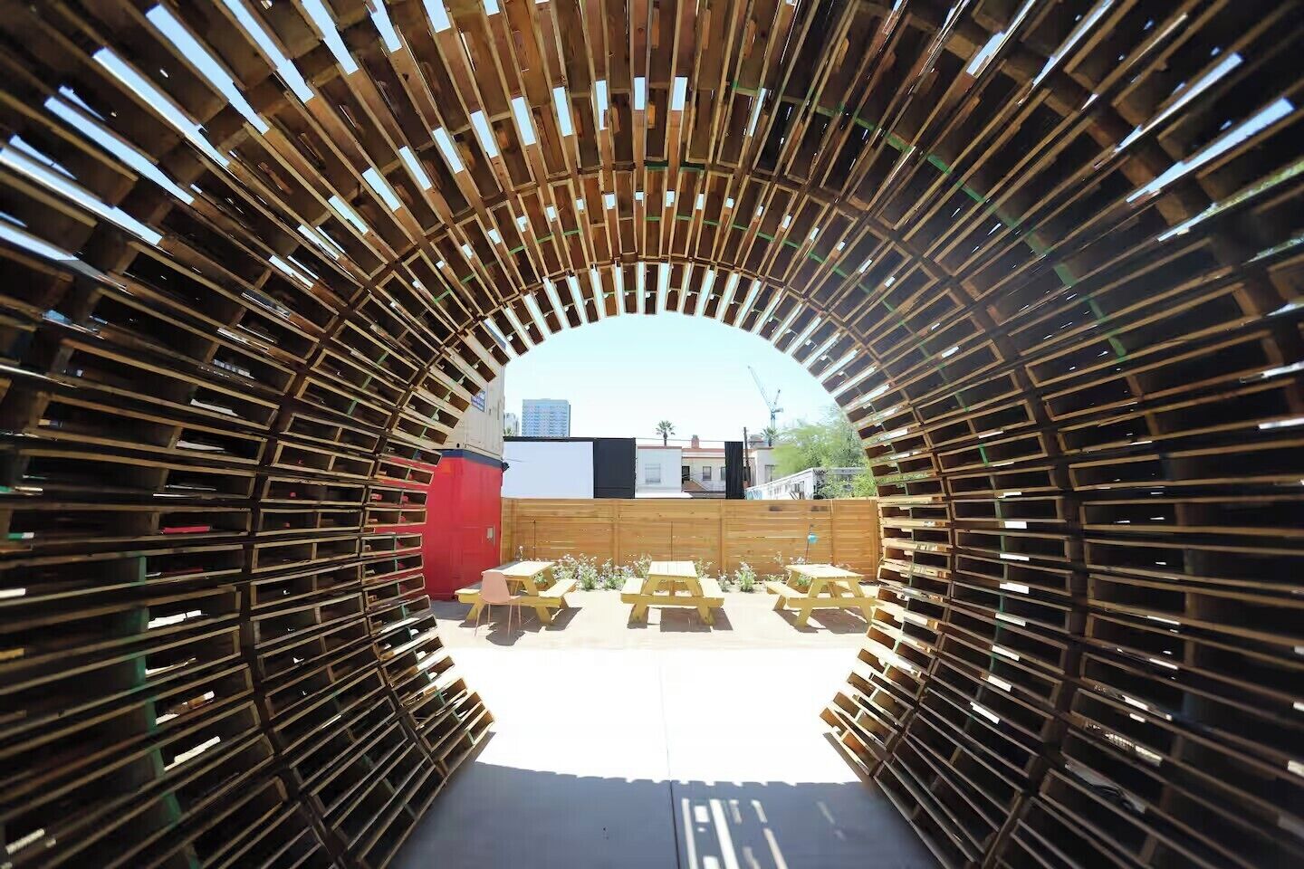 An arched wooden structure with benches and tables, leading to an outdoor area with buildings in the background.