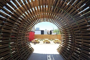 Una estructura de madera arqueada con bancos y mesas, que conduce a un espacio al aire libre con edificios al fondo.