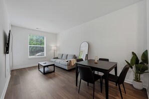 Una sala de estar moderna con un sofá, una mesa de centro, una mesa de comedor con sillas, un televisor montado en la pared y una ventana con persianas.