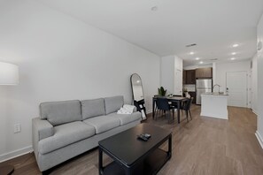 Ein modernes Apartment mit grauem Sofa, einem schwarzen Couchtisch, einem Essbereich mit Tisch und Stühlen sowie einer Küche mit weißen Schränken und einem Kühlschrank.