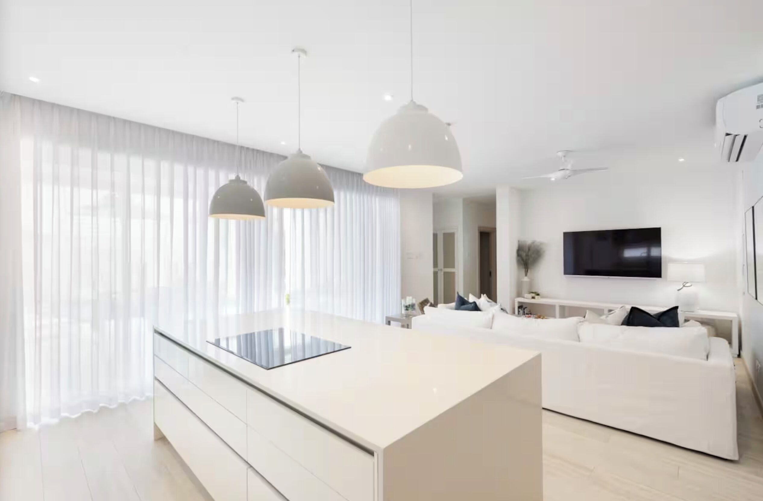 A modern kitchen with a central island, pendant lighting, a white sofa, and a flat-screen TV.