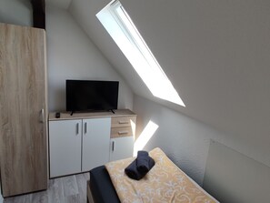 A small room with a slanted ceiling, a television on a wooden cabinet, a bed with a patterned bedspread, and a skylight.