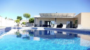 Una piscina con superficie de agua azul cristalina, un porche cubierto con mobiliario exterior, y un edificio de techo plano y amplias ventanas.
