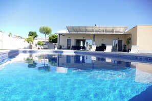 A swimming pool with a clear blue water surface, a covered patio area with outdoor seating, and a building with a flat roof and large windows. - Villa Mi Atardecer (Torrevieja)