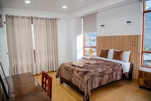 A bedroom with a bed, a wooden table, a chair, a window with curtains, and a view of the outside.