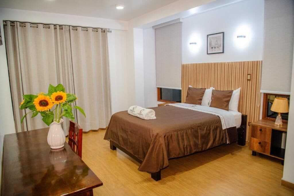 A hotel room with a bed, a bedside table, a lamp, a picture on the wall, and a window with curtains.