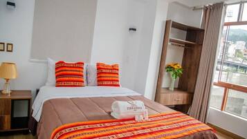 A hotel room with a bed, a nightstand, a wooden wardrobe, a window with a view, and a vase with flowers.