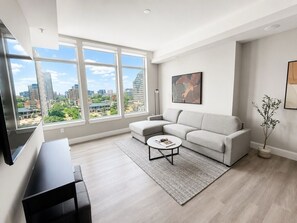 A modern living room with a sectional sofa, a coffee table, and a large window offering a city view. - Level Dallas Uptown (Dallas)