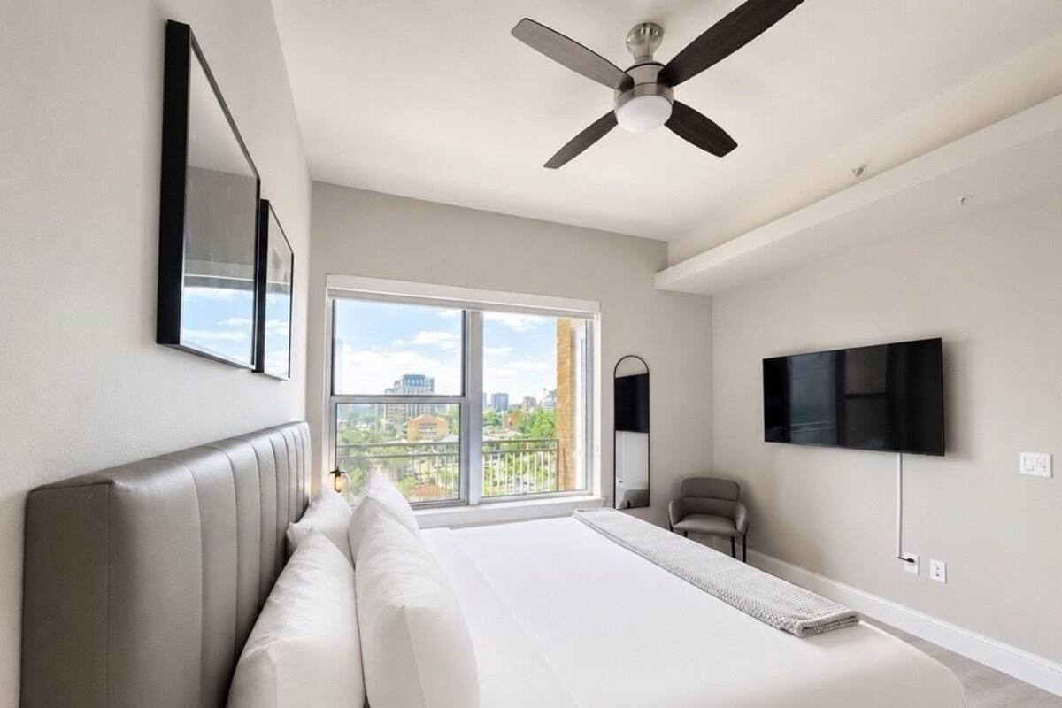 A bedroom with a large bed, a ceiling fan, a TV, and a window with a city view.