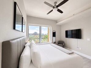 A bedroom with a large bed, a ceiling fan, a TV, and a window with a city view. - Level Dallas Uptown (Dallas)