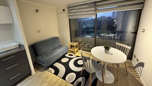 Un salon moderne comprenant un canapé, une table à manger avec des chaises, une petite table d’appoint et une grande fenêtre donnant sur la ville.