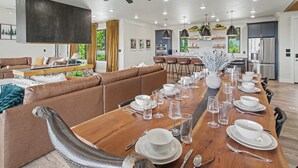 Un comedor moderno con una larga mesa de madera dispuesta para una comida, un sofá de cuero marrón y una cocina al fondo.