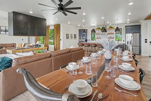 A modern dining area with a long wooden table set for a meal, a brown leather sofa, and a kitchen in the background. - Luxe Group Retreat | Pool, Theater, Views, Games (Sevierville)