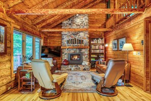 A log cabin interior with a stone fireplace, leather chairs, a bookshelf, and a television.