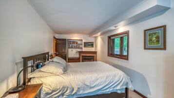 A bedroom with a bed, bedside lamp, bedside table, bookshelf, fireplace, and framed pictures on the wall.