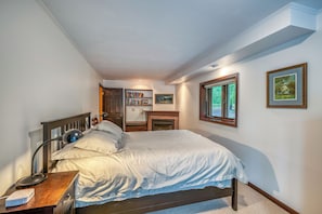 Un dormitorio con cama, lámpara de noche, mesita de noche, estantería, chimenea y cuadros en la pared.