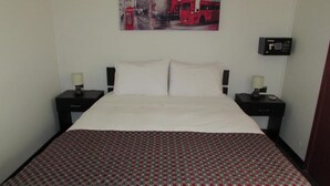 A hotel room with a bed, two bedside tables, a wall-mounted alarm clock, and a cityscape painting above the bed. - Hotel Central (San Fernando)