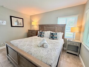 A bedroom with a large bed, two bedside tables with lamps, a headboard, and a window with blinds.