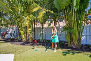 A person playing golf in a backyard with a white fence, palm trees, and a green lawn.
