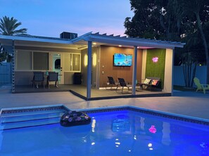 A modern outdoor pool area with a covered seating area, a television, and a neon sign.