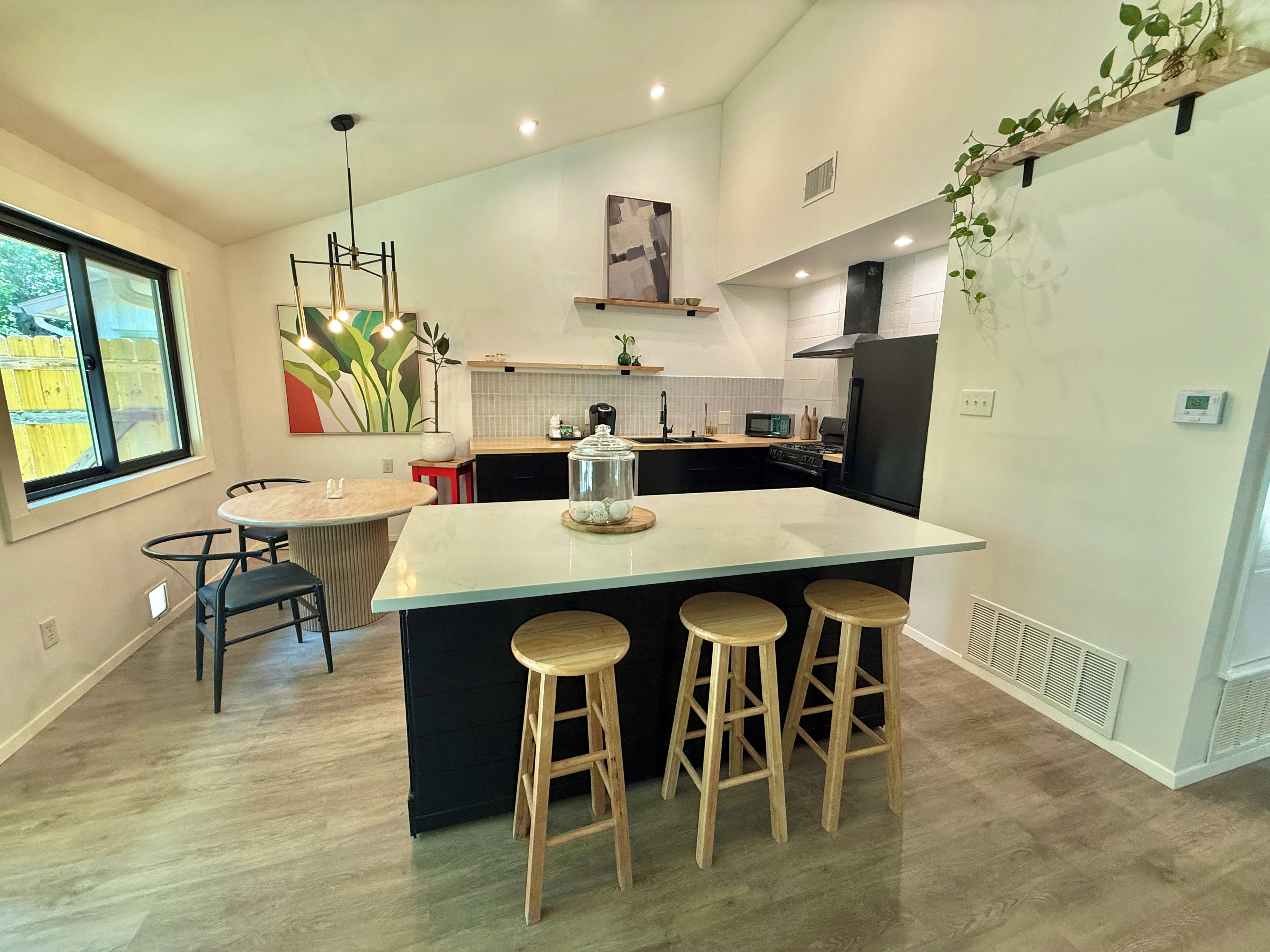 A modern kitchen with a central island, bar stools, a dining table, and a view of the outdoors.