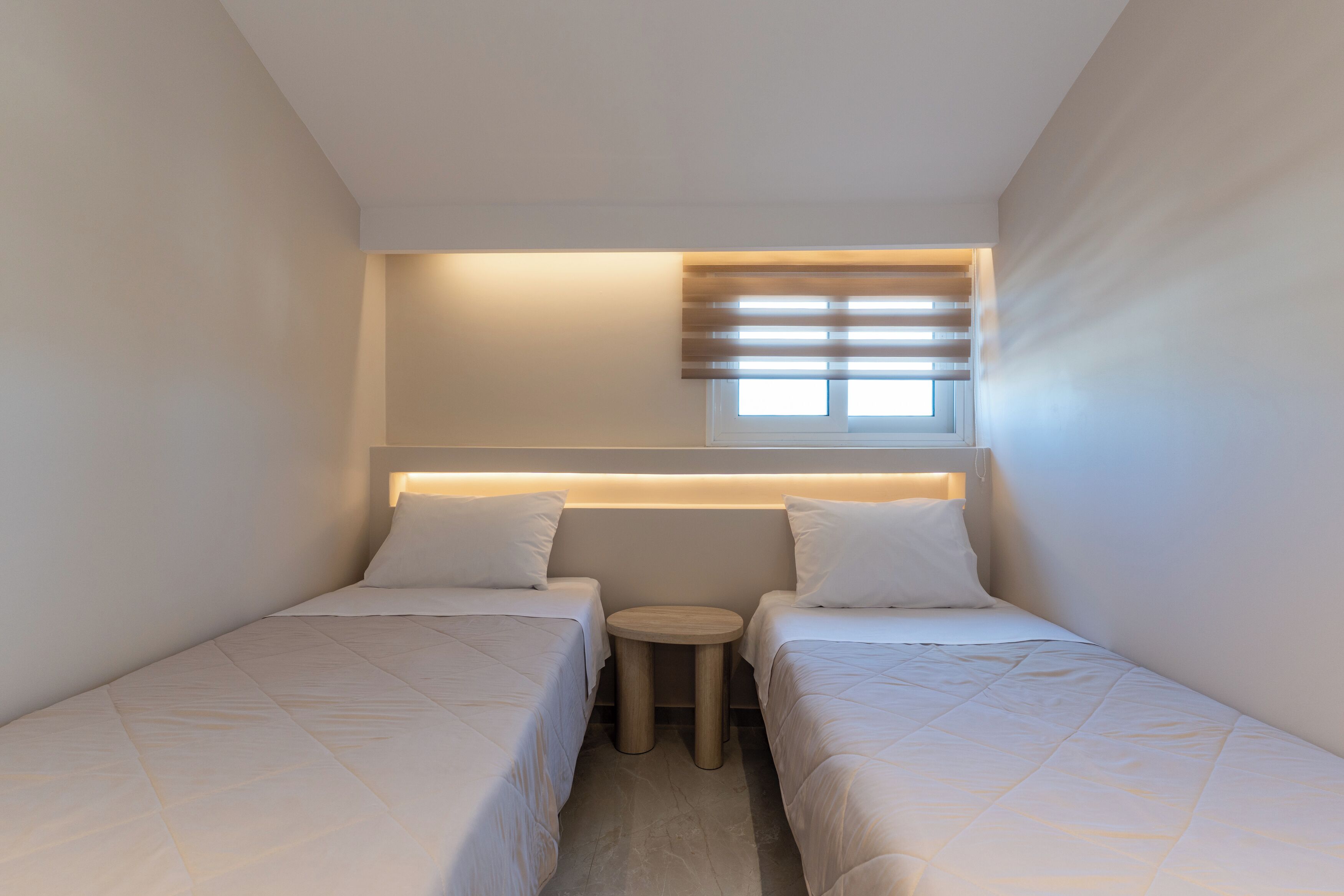 Quarto pequeno e moderno com duas camas, uma mesinha redonda e uma janela com persianas.