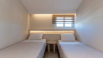 Quarto pequeno e moderno com duas camas, uma mesinha redonda e uma janela com persianas.