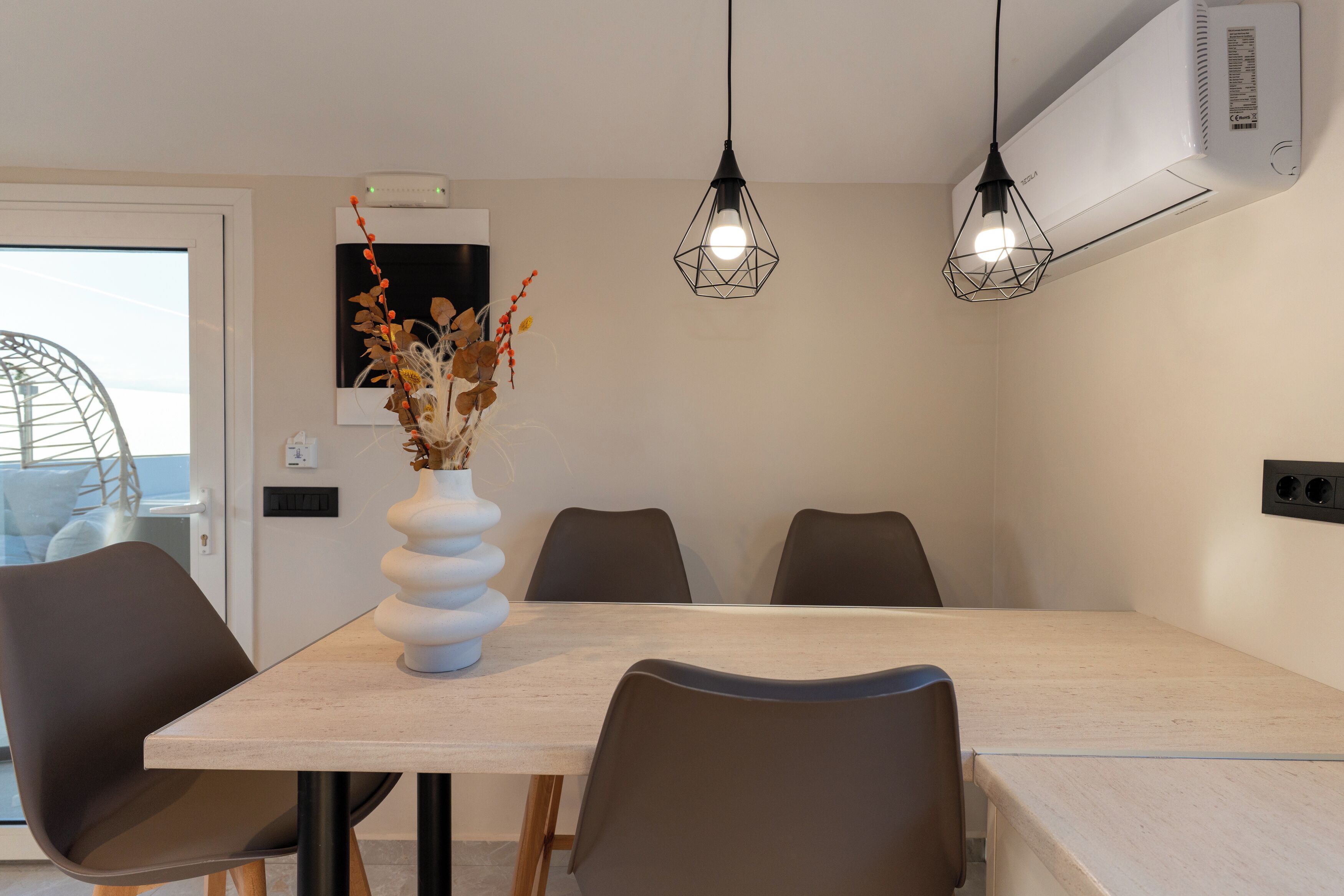 A modern dining area with a light-colored table, dark chairs, a white vase with dried flowers, and two hanging pendant lights.