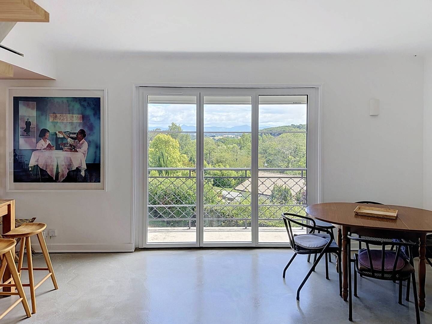 A room with a large sliding glass door leading to a balcony with a view of trees and buildings. There is a framed picture on the wall, a round table with chairs, and wooden stools.
