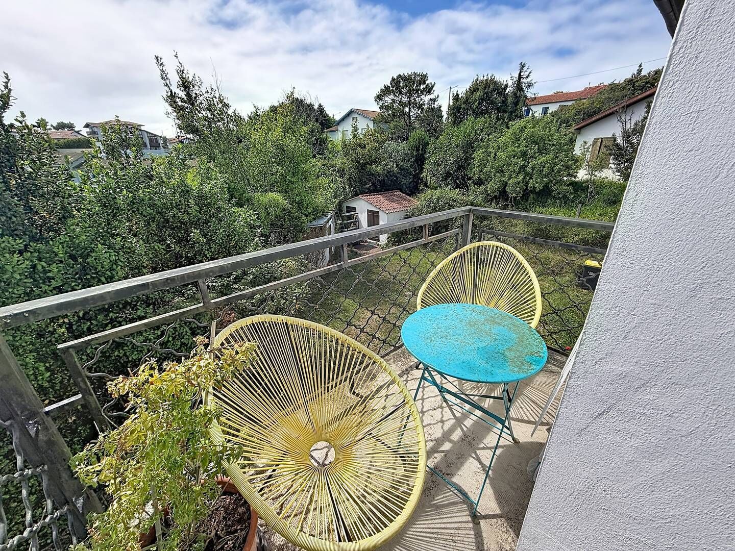 Un balcon avec deux chaises et une table, entouré de verdure et avec des maisons en arrière-plan.