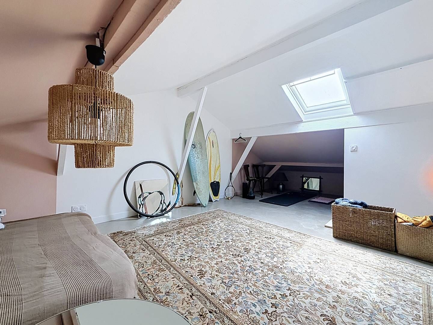 A spacious loft bedroom with a large bed, a hanging light fixture, a skylight, and surfboards against the wall.