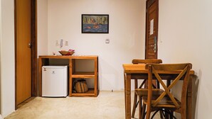 A compact kitchen with a refrigerator, wooden table, and chairs, a small shelf with a bowl, and a framed picture on the wall.