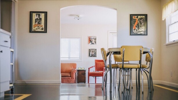 A hallway with a dining area featuring a yellow chair, a red chair, and a small table. There are framed pictures on the walls and a window with blinds.