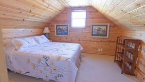 A bedroom with a wooden sloped ceiling, a bed with floral bedding, two framed pictures on the wall, and a small wooden table.
