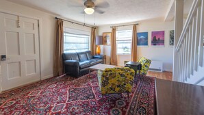 A living room with a black sofa, a yellow armchair, a patterned rug, a ceiling fan, and a window with curtains.
