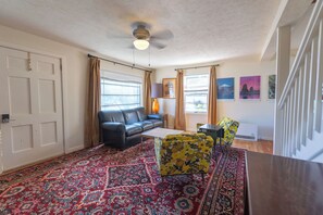 Un salon avec un canapé noir, un fauteuil jaune, un tapis à motifs, un ventilateur de plafond et une fenêtre avec des rideaux.