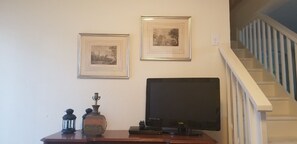 A room with a flat-screen TV on a wooden stand, two framed pictures on the wall, a small table with a lantern and a vase, and a staircase with white railings.