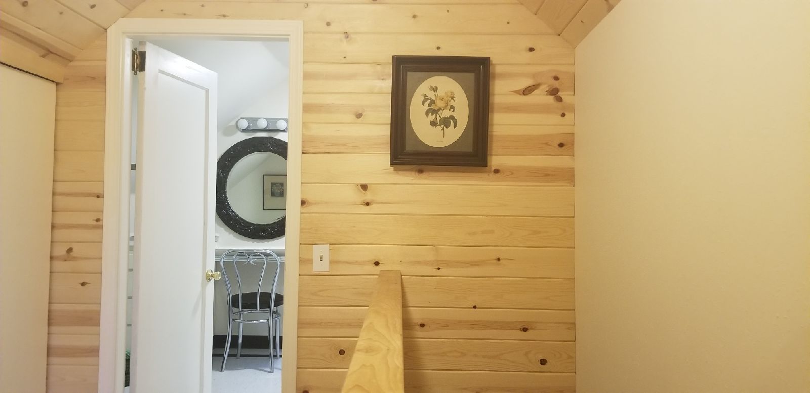A wooden interior with a door leading to another room, a framed picture on the wall, and a mirror above a chair.