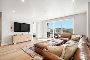 Een ruime woonkamer met een grote leren zithoekbank, een flatscreen televisie aan de muur en een grote glazen schuindeur die leidt naar een terras.