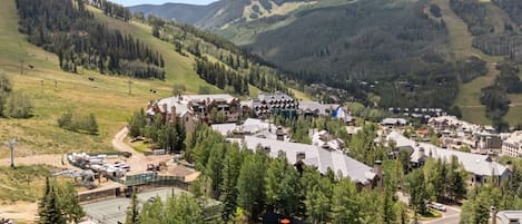 Un complejo turístico de montaña con varias construcciones, una cancha de tenis y colinas boscosas circundantes.