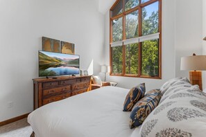 Ein Schlafzimmer mit einem großen Bett, einem Schminktisch, einem Fernseher und einem Fenster mit Blick auf Bäume.