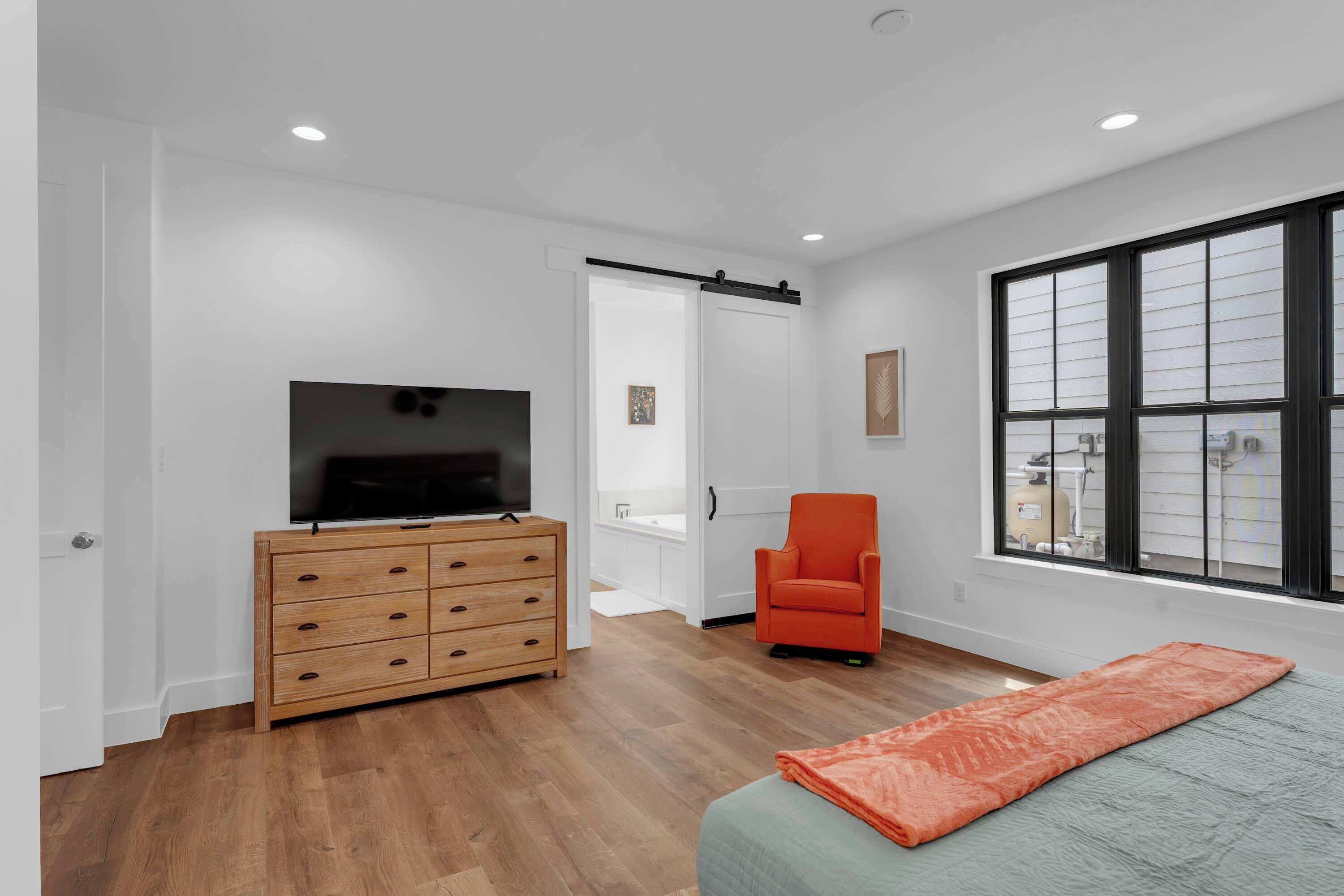 Een slaapkamer met een houten dressoir, een televisie, een oranje fauteuil, een bed met een oranje deken en een groot raam met jaloezieën.