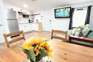 Cocina compacta con armarios blancos, microondas integrado y refrigerador. Hay una mesa de comedor con un centro de mesa de girasol.