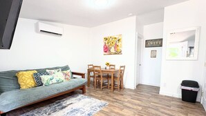 Uma sala de estar com sofá, mesa de jantar com cadeiras, um quadro e uma lixeira.