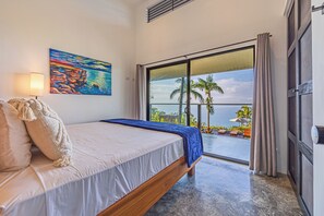 Ein Schlafzimmer mit einem Bett, einem Gemälde, einer Lampe und Blick auf den Ozean durch eine Schiebetür.