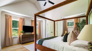Un lit à baldaquin avec un téléviseur posé sur une commode en bois, une grande fenêtre avec des rideaux et un ventilateur de plafond.