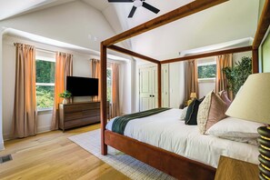 Ein Himmelbett mit Fernseher auf einer Holzkommode, ein großes Fenster mit Vorhängen und ein Deckenventilator.