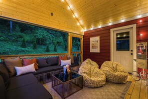 Ein gemütliches Interieur mit Holzbalkendecke, einer Couch, Sitzsäcken und einem großen Fenster mit Blick auf den Wald.