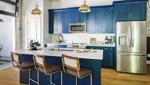 Une cuisine moderne avec des armoires bleues, des appareils en acier inoxydable, un îlot central doté de tabourets de bar, un éclairage suspendu et une vue sur l’extérieur.