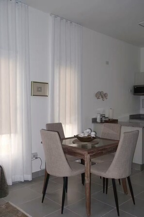 Un coin repas avec une table en bois et quatre chaises, un coin cuisine équipé d’un four à micro-ondes et un rideau blanc.
